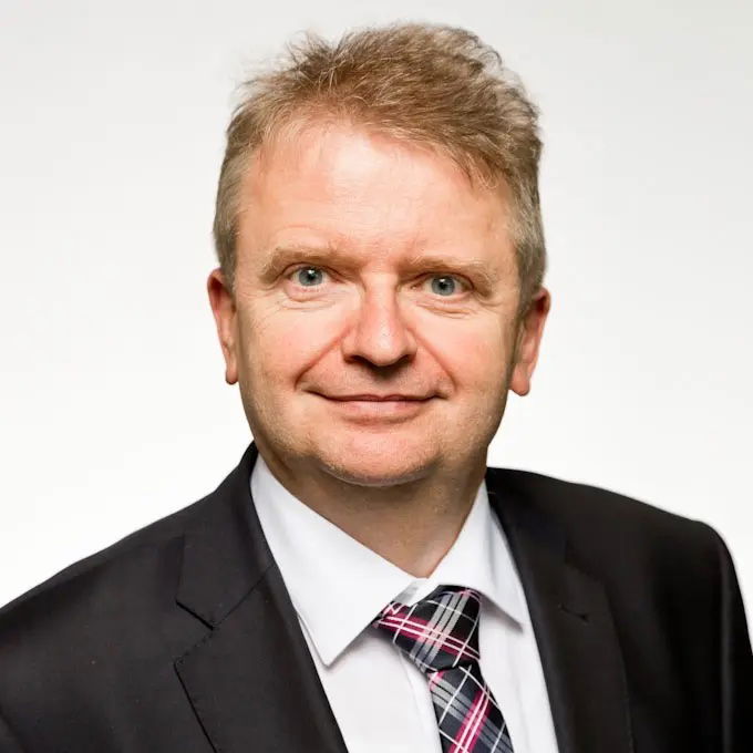 Porträtfoto des Investor Redakteurs Dimitri Speck im Anzug mit weißem Hemd und kariertem Schlips vor hellem Hintergrund. Der Mann hat helles Haar, blaue Augen und lächelt leicht in die Kamera.